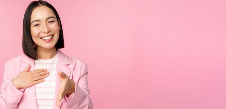 Portrait of smiling, pleasant businesswoman shaking hands with business partner, handshake, extending hand and saying hello, standing over pink background