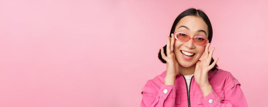 Cute modern japanese girl in sunglasses, smiling and looking happy, posing against pink background in stylish clothing