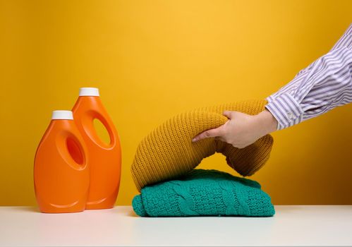 washed folded clothes and plastic orange large bottles with liquid detergent stand on a white table, yellow background. Routine homework