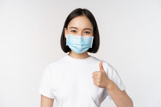 Smiling asian girl in medical mask showing thumbs up, approve something good, praise and compliment company, standing over white background