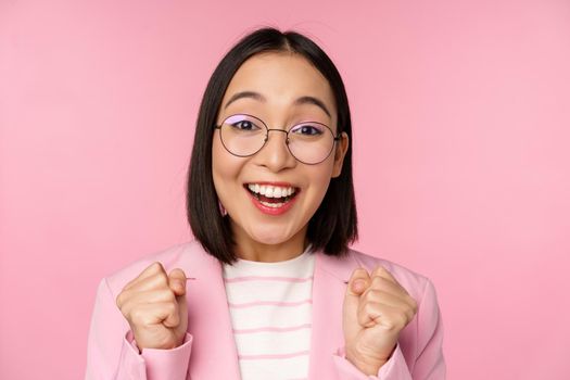 Close up portrait of asian businesswoman cheering, rooting for, looking with hope and excitement at camera, smiling and laughing, standing over pink background