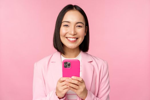Portrait of smiling business woman, asian corporate person using smartphone, mobile phone application, standing over pink background