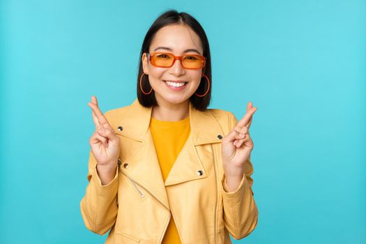 Smiling beautiful asian woman wishing, cross fingers for good luck and looking hopeful, standing over blue background