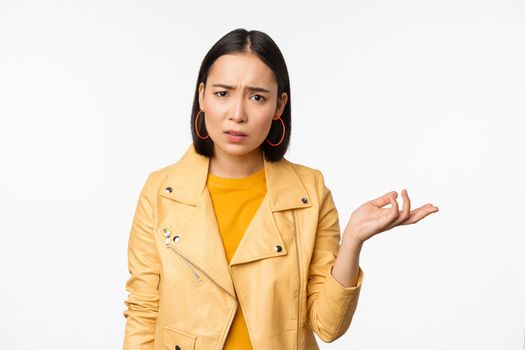 Image of frustrated korean woman, shrugging shoulders and raising hand, looking puzzled, cant understand smth, standing over white background