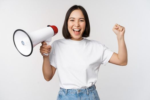 Happy asian woman shouting at megaphone, making announcement, advertising something, standing over white background