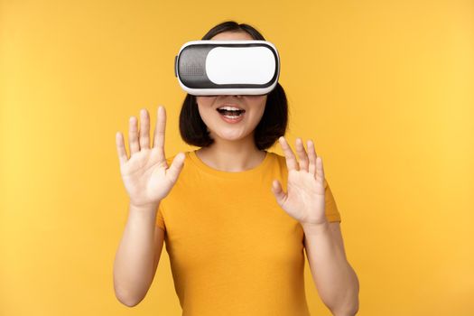 Young asian woman playing in VR, using virtual reality glasses, wearing yellow t-shirt against studio background