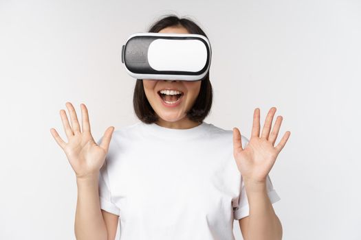 Happy asian woman using VR headset, waving raised hands and laughing, using virtual reality glasses, standing over white background
