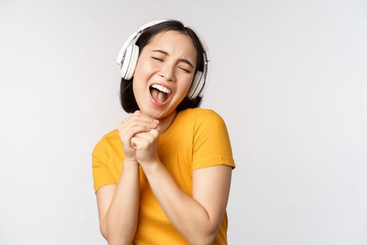 Happy asian girl dancing, listening music on headphones and smiling, standing in yellow tshirt against white background