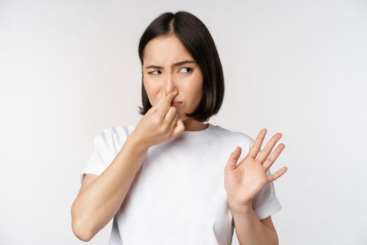 Asian girl looks disgusted, rejecting product with bad smell, shut nose from aversion and cringe, standing against white background