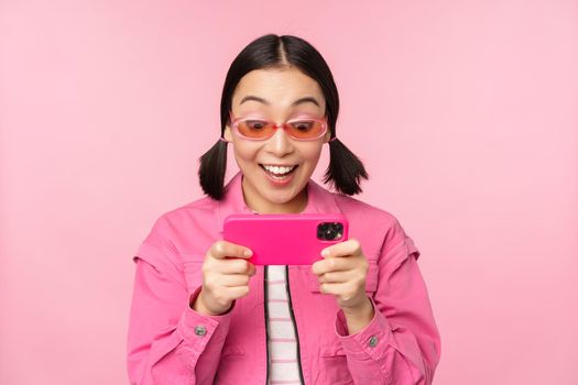 Portrait of happy asian girl playing on smartphone, watching videos on mobile phone app, standing over pink background