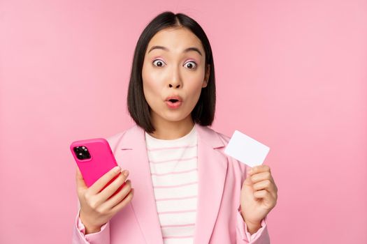 Amazed businesswoman, asian girl in suit showing credit card and mobile phone, order online, shopping with smartphone, standing over pink background. Advertising concept