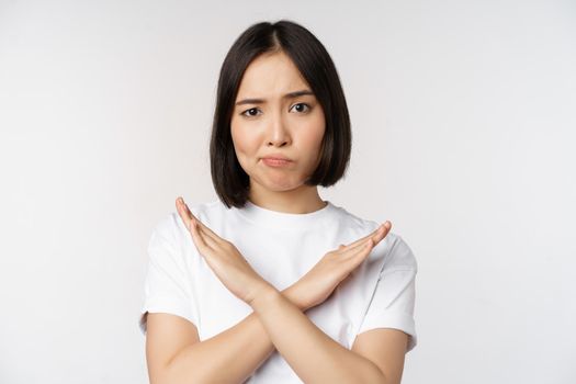 Portrait of asian korean woman showing stop, prohibition gesture, showing arm cross sign, standing in tshirt over white background