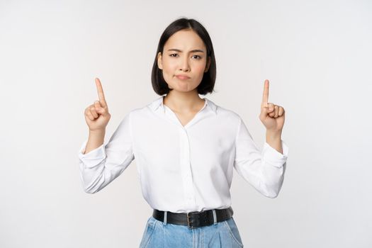 Image of skeptical asian lady grimacing, frowning upset, pointing fingers up, showing advertisement, standing over white background