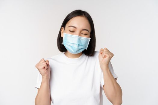 Covid-19, healthcare and medical concept. Enthusiastic asian woman in medical face mask, dancing and celebrating, winning, achieve goal, standing over white background