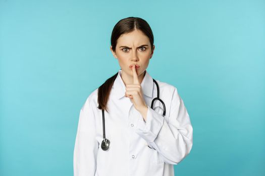 Angry female doctor, healthcare medical worker shushing with disapproval, taboo quiet gesture, silence someone, standing over torquoise background