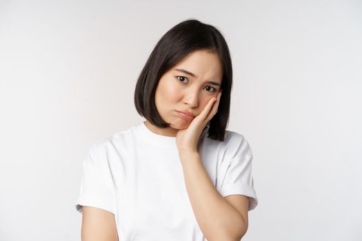 Sad asian girl looking upset and lonely, sulking and frowning, standing against white background in casual tshirt