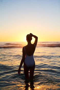 We all get lost in the beauty sometimes. Rearview shot of a young woman standing in the water at sunset.