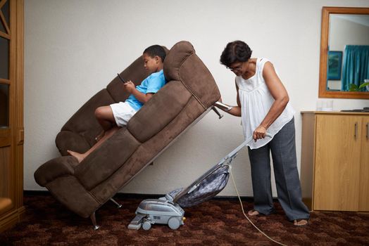 Grandmom is a superwoman. Shot of a grandmother vacuuming under a couch which her grandson is lying on.