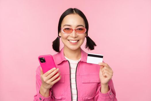 Online shopping. Smiling asian girl shopper, holding smartphone and credit card, paying in mobile app, standing over pink background