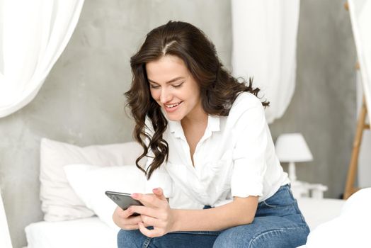Happy young woman with mobile phone sitting on bed and browsing social media at home.