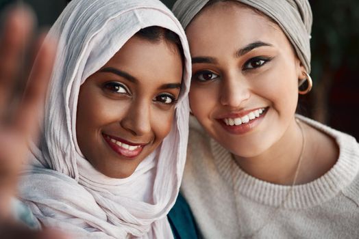 Capture this. Cropped portrait of two affectionate young girlfriends taking a selfie together at a cafe while dressed in hijab.