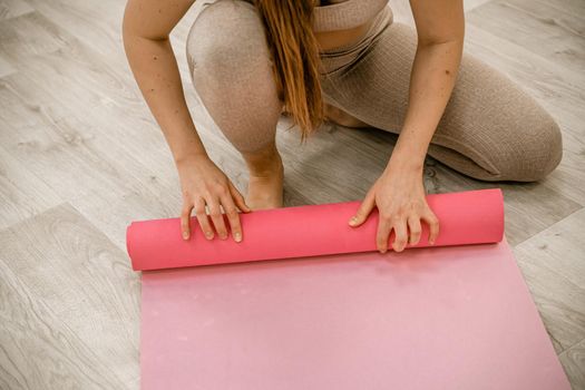 A young woman rolls a pink fitness or yoga mat before or after exercising, exercising at home in the living room or in a yoga studio. Healthy habits, keep fit, weight loss concept. Closeup photo