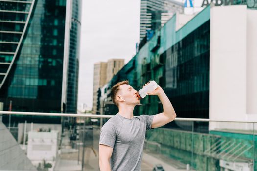 Tired Young man runner drinking water, relaxing after sport training. Holding water bottle while doing fitness workout in summer city urban street, cloudy sky