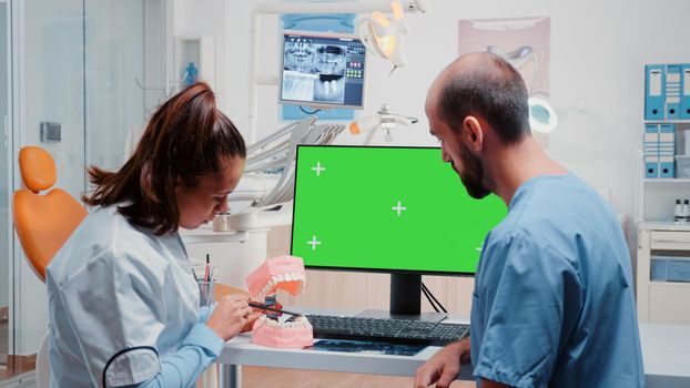 Oral care team analyzing teeth layout while using computer with horizontal green screen on desk. Dentist and dental assistant working with tools and equipment for dentistry and teethcare