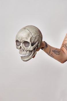Tattooed hand of a woman in a black watch is holding a realistic model of a human skull with teeth isolated on white. Close-up shot.