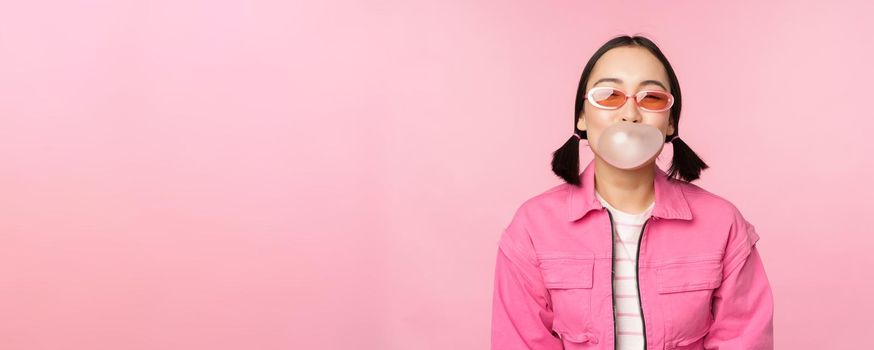 Stylish asian girl blowing bubblegum bubble, chewing gum, wearing sunglasses, posing against pink background