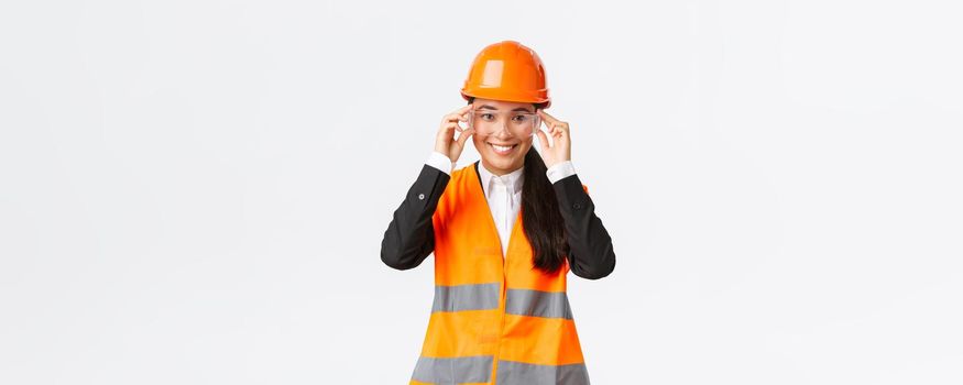 Smiling pretty female asian engineer, construction manager in safety helmet and glasses following protocol before enterting building area or enterprise, standing white background