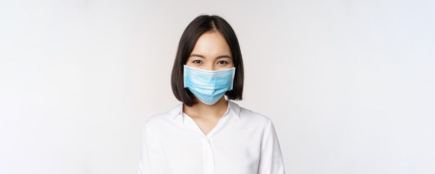 Covid and healthcare concept. Close up portrait of asian woman, office lady in face mask, smiling, using protection from coronavirus during pandemic, white background.