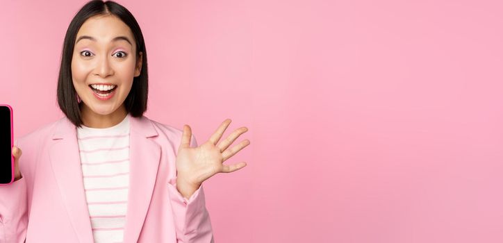 Surprised, enthusiastic asian businesswoman showing mobile phone screen, smartphone app interface, standing against pink background