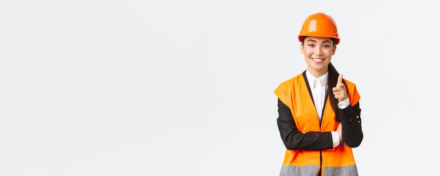 Successful and confident smiling asian female architect pointing finger at camera, wearing safety helmet and reflective jacket, introduce construction project, industrial woman inviting clients