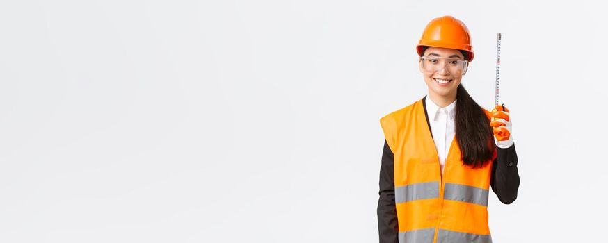 Confident smiling, professional asian female engineer, construction technician in safety helmet and reflective uniform, standing with tape measure, measuring layout at building area