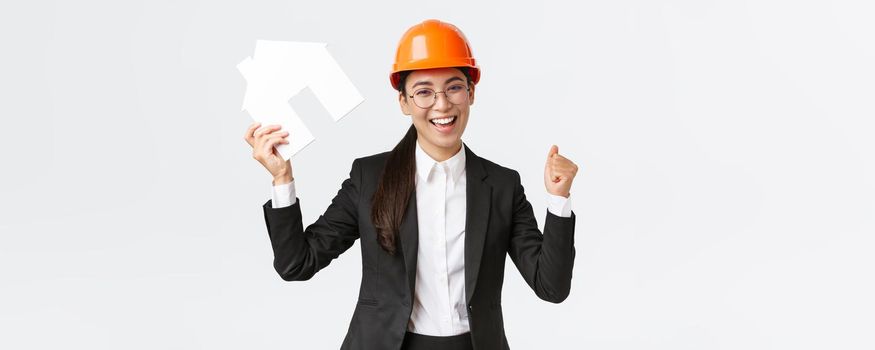 Successful winning asian female construction engineer, team lead architect achieve goal, holding house maket and fist pump in rejoice, celebrating victory in helmet and business suit