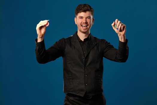 Newbie in poker, in black vest and shirt. Holding some colored chips. Posing against blue background. Gambling, casino. Close-up.