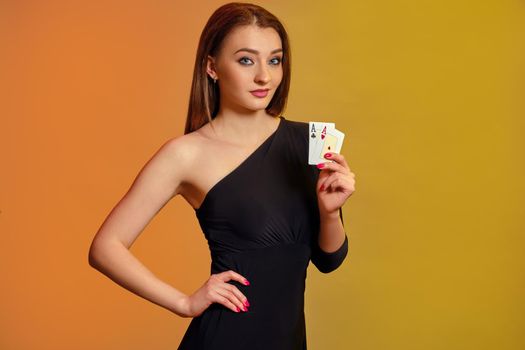 Blonde model with bright make-up, in black dress is showing two aces, posing against colorful background. Gambling, poker, casino. Close-up.