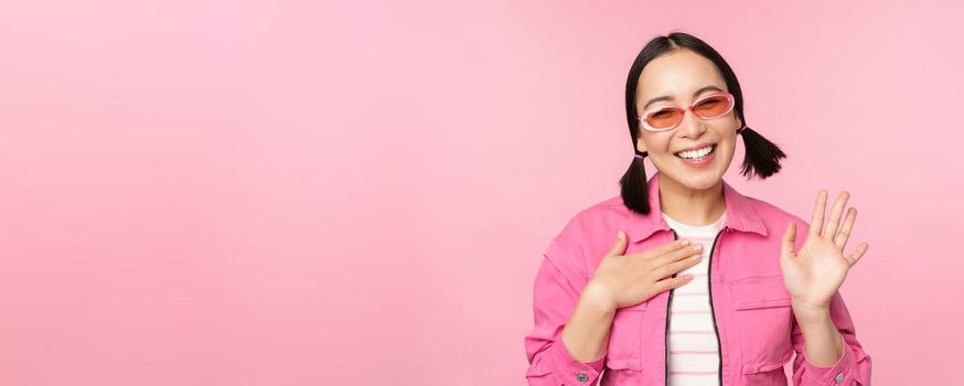 Portrait of friendly asian teen girl in sunglasses saying hello, waves her hand and smiles, greets you, hi gesture, stands over pink background