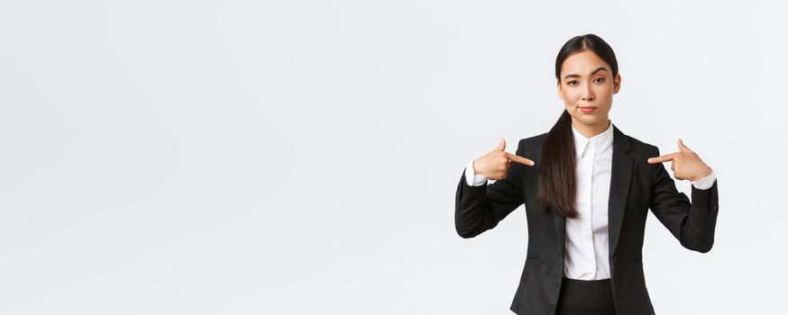 Professional asian businesswoman in suit pointing at herself as offering best deals. Proud and confident saleswoman show-off, suggest personal help or assistance, standing white background