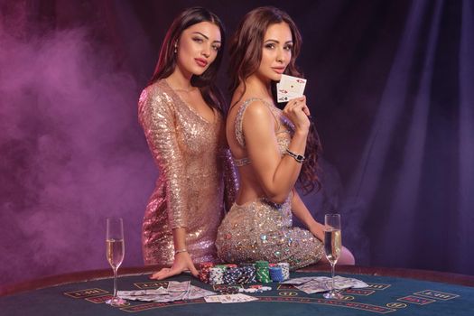 Two gorgeous women showing cards while posing at playing table in casino. Black, smoke background with colorful backlights. Gambling, poker. Close-up.
