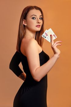 Blonde girl with bright make-up, in black dress is showing two aces, posing sideways against colorful background. Gambling, poker, casino. Close-up.