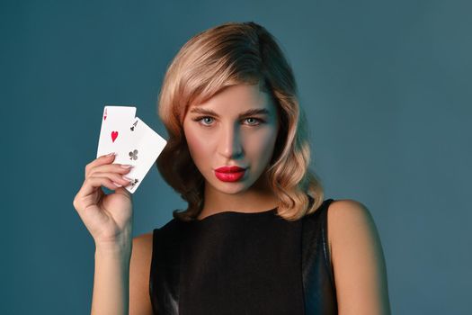 Blonde girl in black stylish dress showing two playing cards, posing against blue background. Gambling entertainment, poker, casino. Close-up.