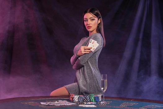 Woman in gray dress showing cards, sitting on playing table in casino. Black, smoke background with colorful backlights. Gambling, poker. Close-up.