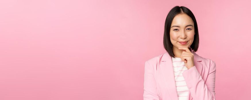 Image of asian businesswoman standing in thinking pose, brainstoming, wearing suit. Korean saleswoman, entrepreneur posing against pink background