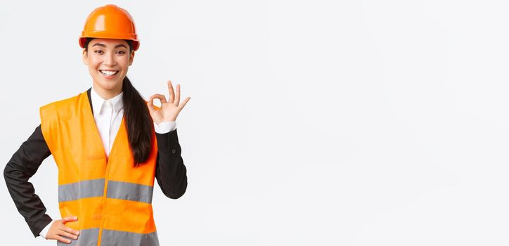 Confident professional asian female architect in safety helmet assure quality and work in time, showing okay gesture and smiling determined, standing assertive, ensure and guarantee something