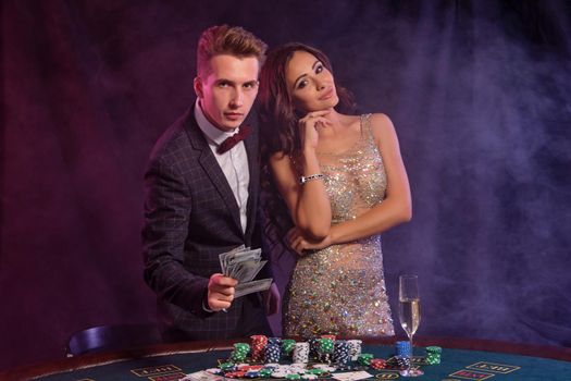 Man and woman playing poker at casino, celebrating win at table with stacks of chips, money, cards, champagne. Black, smoke background. Close-up.