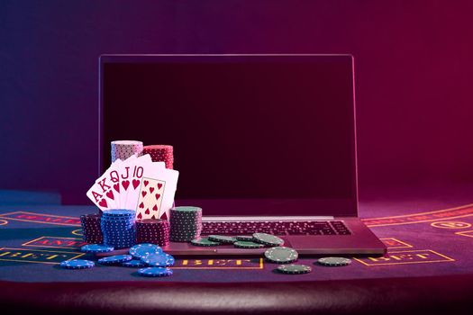 Chips piles and playing cards on laptop standing on blue cover of a table. Black background. Gambling entertainment, poker, casino concept. Close-up.