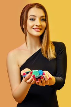 Blonde lady in black dress is smiling, showing handful of multicolored chips, posing on colorful background. Poker, casino. Close-up.