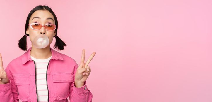 Beautiful korean girl in sunglasses, blowing bubblegum bubble and showing peace signs, standing over pink background